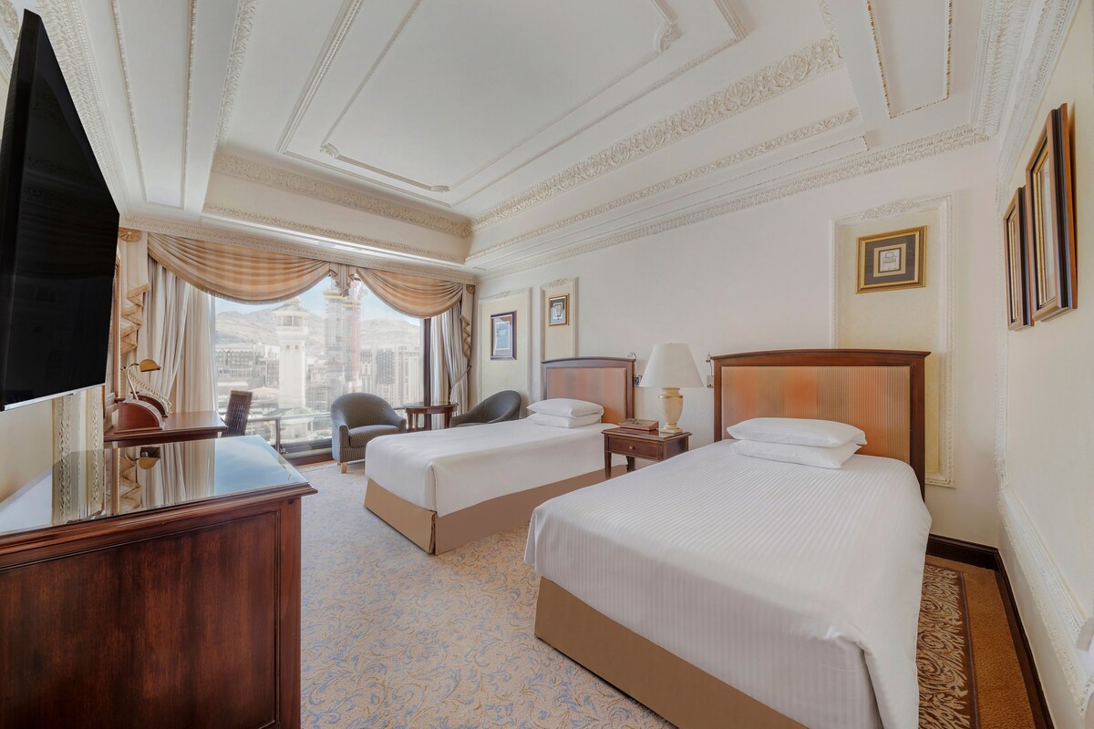 A hotel room with two beds, a desk, a chair, a TV, and a view of a cityscape.