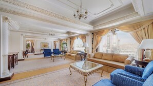 Un salon spacieux avec un grand canapé, une table basse et un fauteuil bleu. Un lustre est suspendu au plafond et de grandes fenêtres offrent une vue sur le paysage urbain.