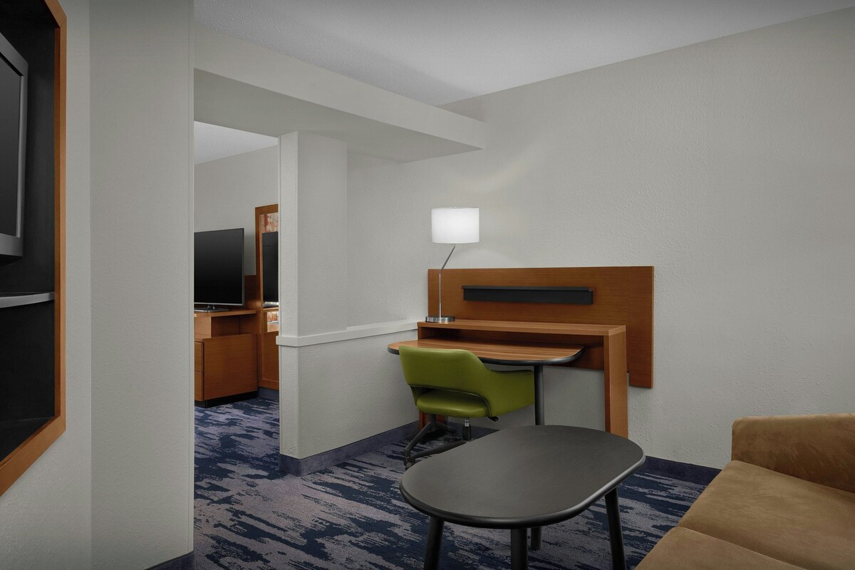 A compact hotel room with a kitchenette, a single bed, a desk with a chair, a small table, and a sofa.