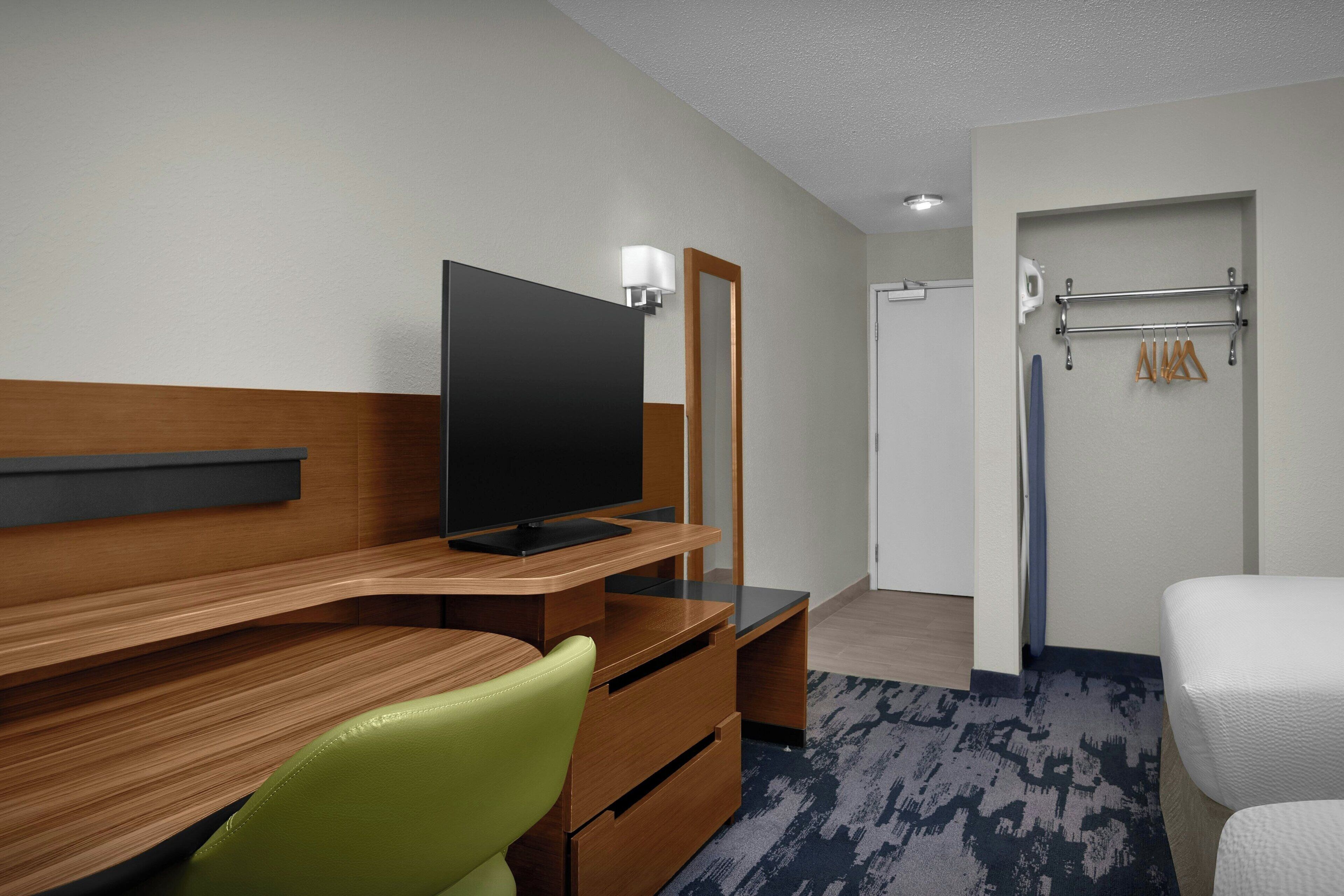 Habitación de hotel con un escritorio de madera, una silla verde, un televisor de pantalla plana, un espejo, una cama blanca y un perchero para toallas.