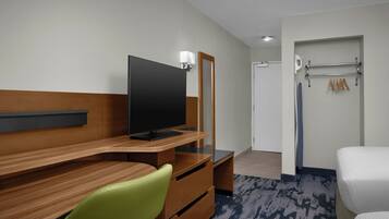 Habitación de hotel con un escritorio de madera, una silla verde, un televisor de pantalla plana, un espejo, una cama blanca y un perchero para toallas.