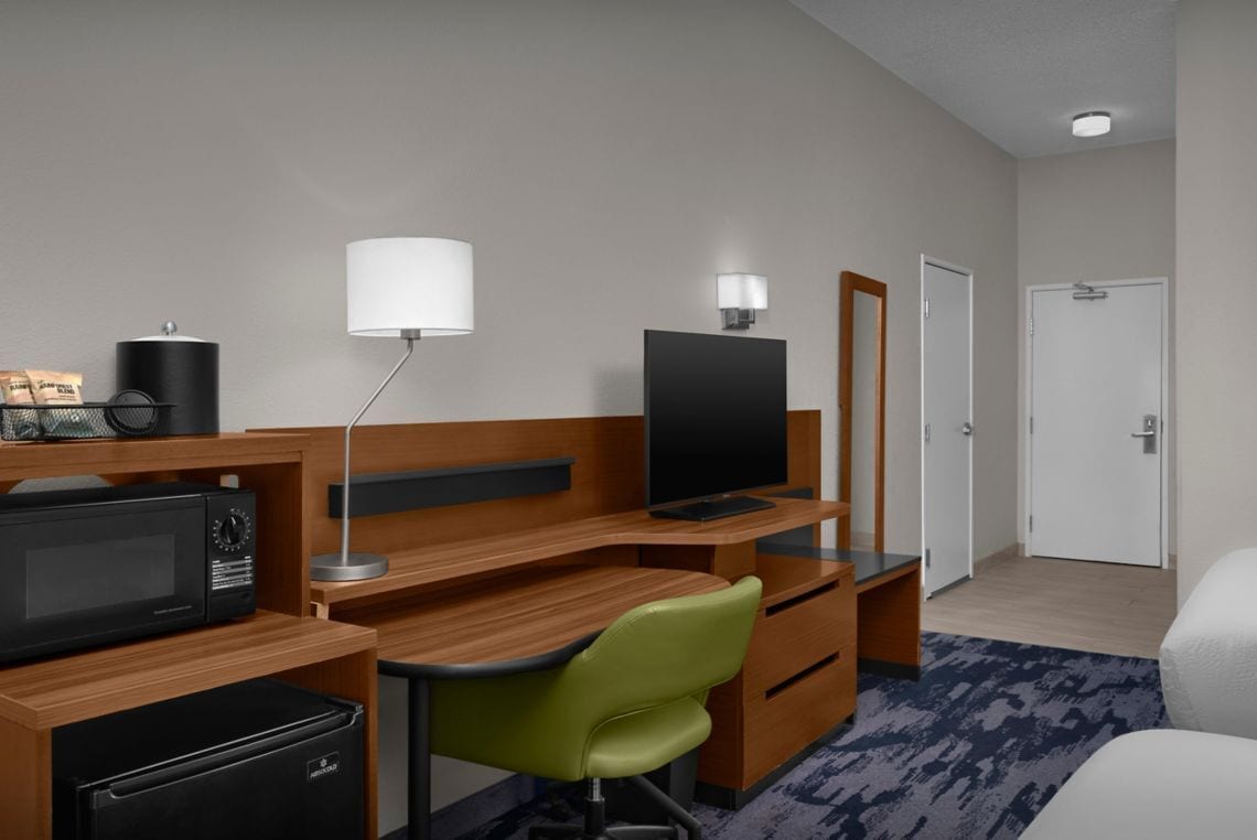 A hotel room with a wooden desk, a green chair, a television, a microwave, a refrigerator, a lamp, and a door.