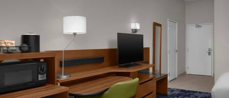 A hotel room with a wooden desk, a green chair, a television, a microwave, a refrigerator, a lamp, and a door.