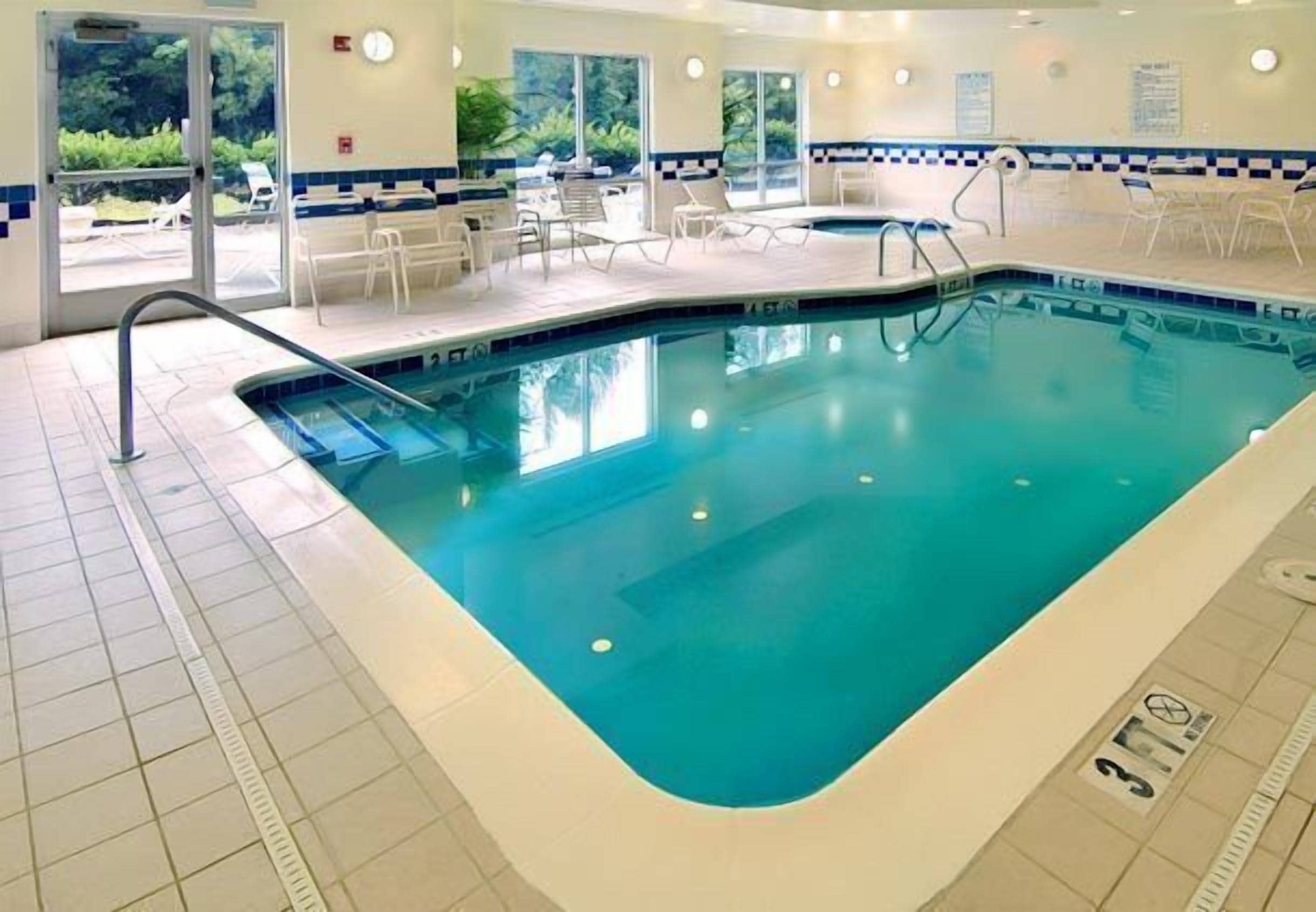 An indoor swimming pool with clear water, a tiled floor, and a poolside area with chairs and a sunshade.