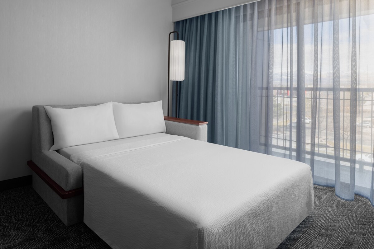 Une chambre d’hôtel avec un lit, une table de chevet, une lampe et une vue sur la ville à travers de fines rideaux.