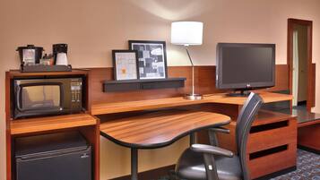 A compact hotel room with a wooden desk, a microwave, a refrigerator, a television, and a chair.