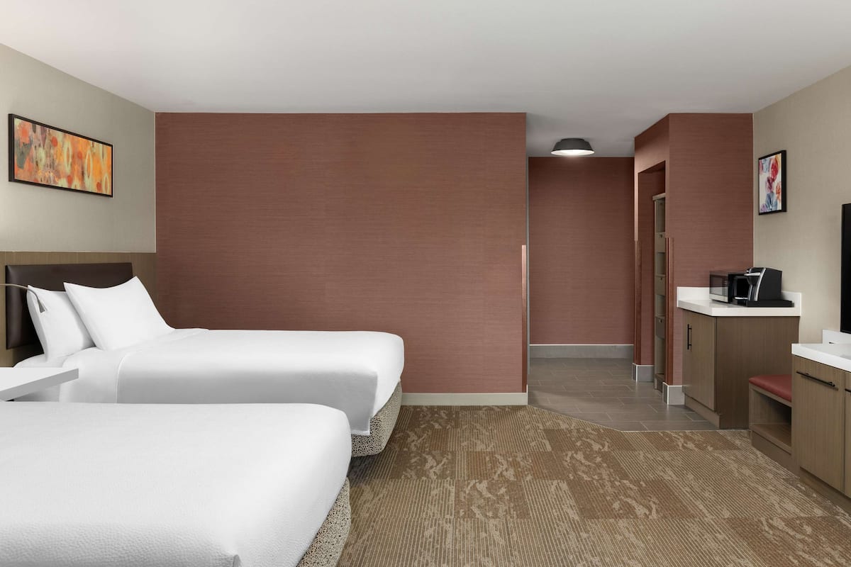 A hotel room with two beds, a kitchenette, a bathroom, and a large window.