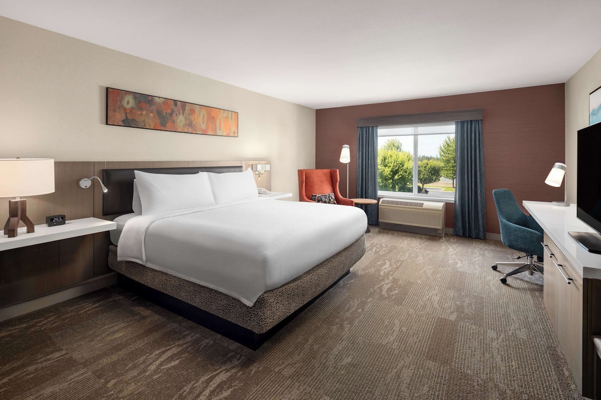 A hotel room with a large bed, a desk with a chair, a window with curtains, and a painting on the wall.