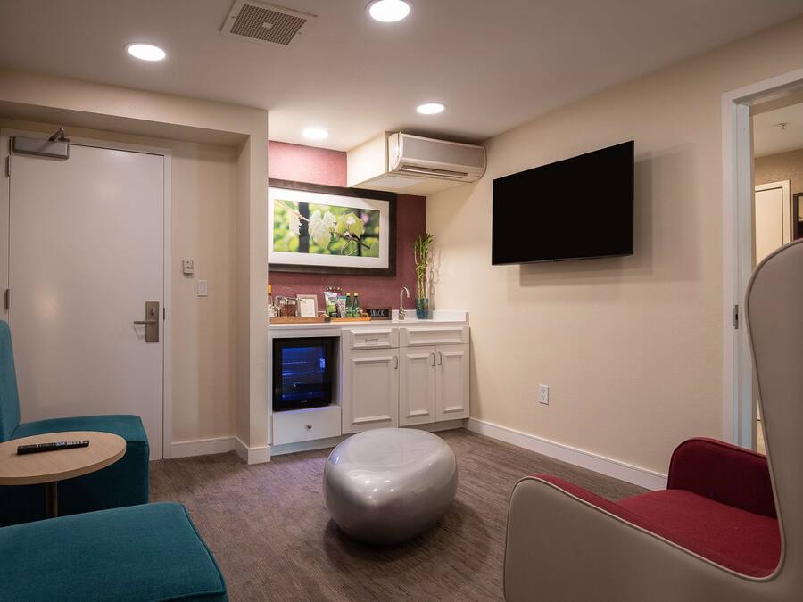 Un espacio habitable compacto con kitchenette, un televisor de pantalla plana montado en la pared, un pouf redondo y una pequeña mesa redonda con un control remoto.