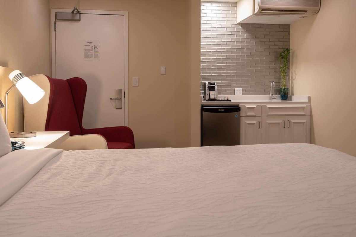 A hotel room with a bed, a desk with a lamp, a red chair, a kitchenette with a sink, and a door with a bulletin board.