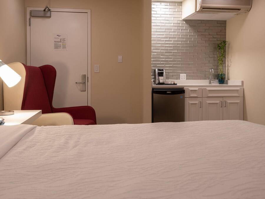 A hotel room with a bed, a desk with a lamp, a red chair, a kitchenette with a sink, and a door with a bulletin board.