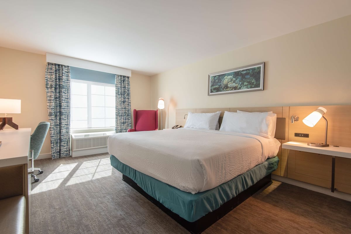 A hotel room with a large bed, a desk with a chair, a red chair, a window with curtains, and a wall-mounted lamp.