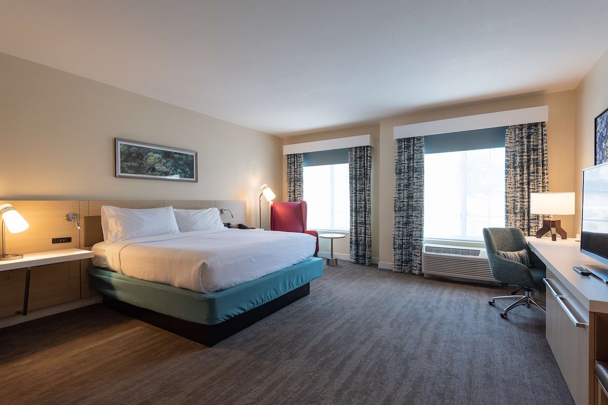 A hotel room with a bed, desk, chair, and a large window with curtains.