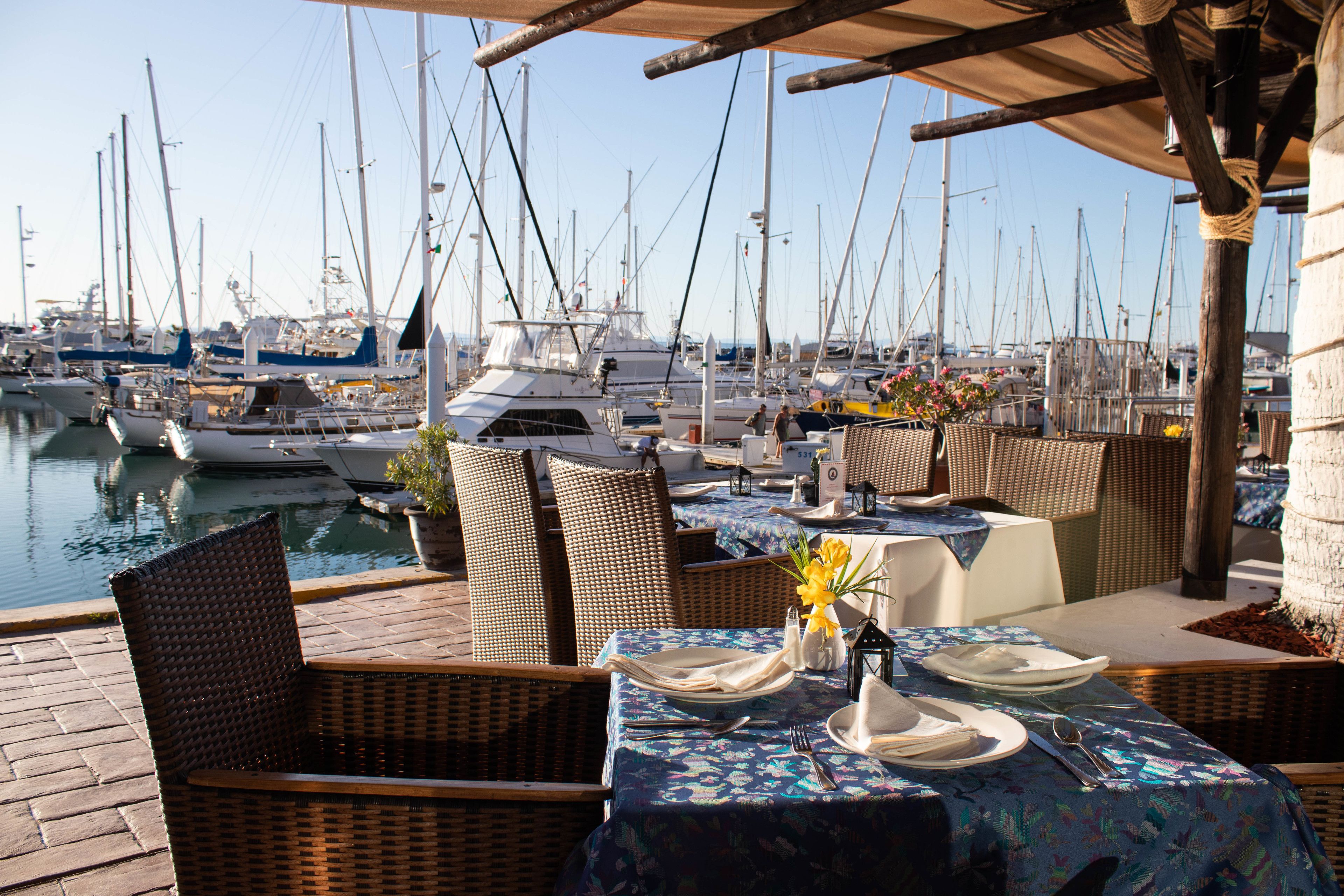 A marina with boats docked, outdoor dining area with wicker chairs and tables set for two, and a view of the water.