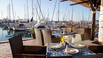 A marina with boats docked, outdoor dining area with wicker chairs and tables set for two, and a view of the water.