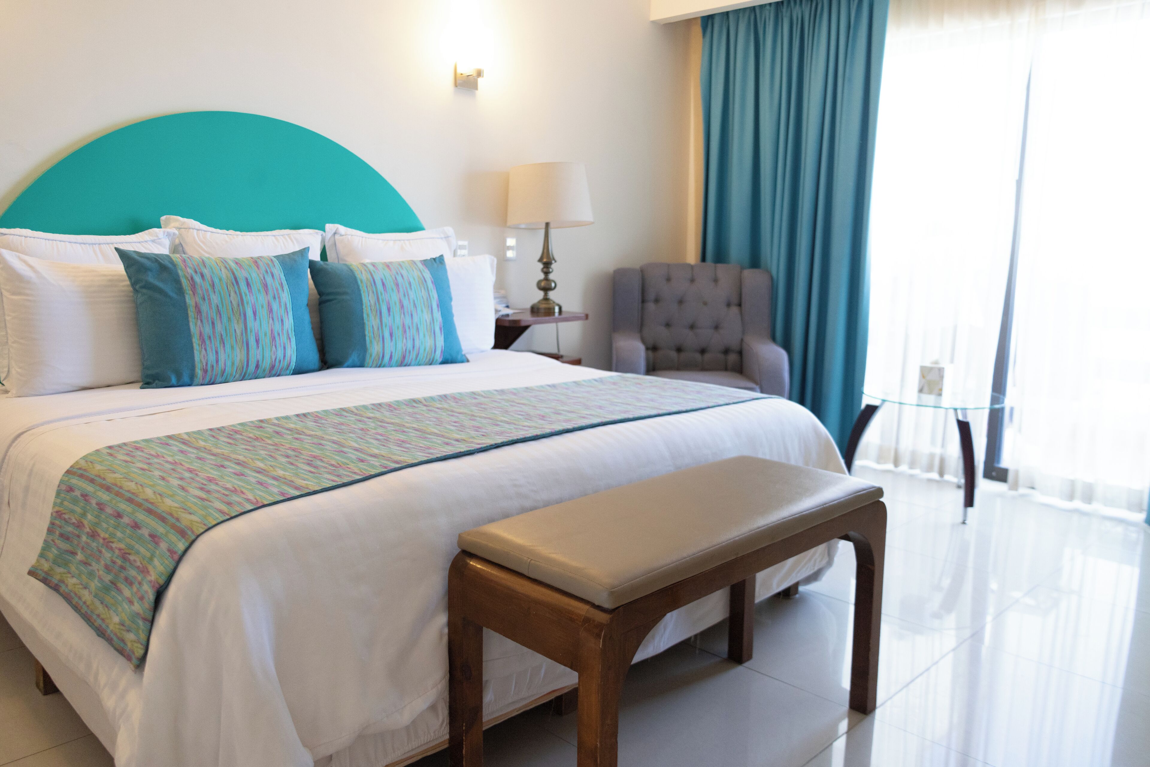 A hotel room with a large bed, a bench, a chair, a lamp, and a view of the outdoors.