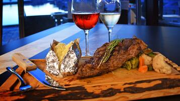 Une planche à découper en bois sur laquelle on voit un steak, des légumes et des aliments emballés dans du papier d’aluminium, le tout accompagné de deux verres de vin, sur une table offrant une vue sur un plan d’eau.