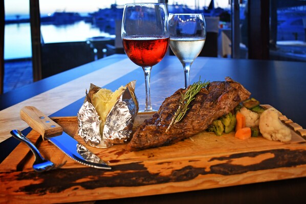 Une planche à découper en bois sur laquelle on voit un steak, des légumes et des aliments emballés dans du papier d’aluminium, le tout accompagné de deux verres de vin, sur une table offrant une vue sur un plan d’eau.