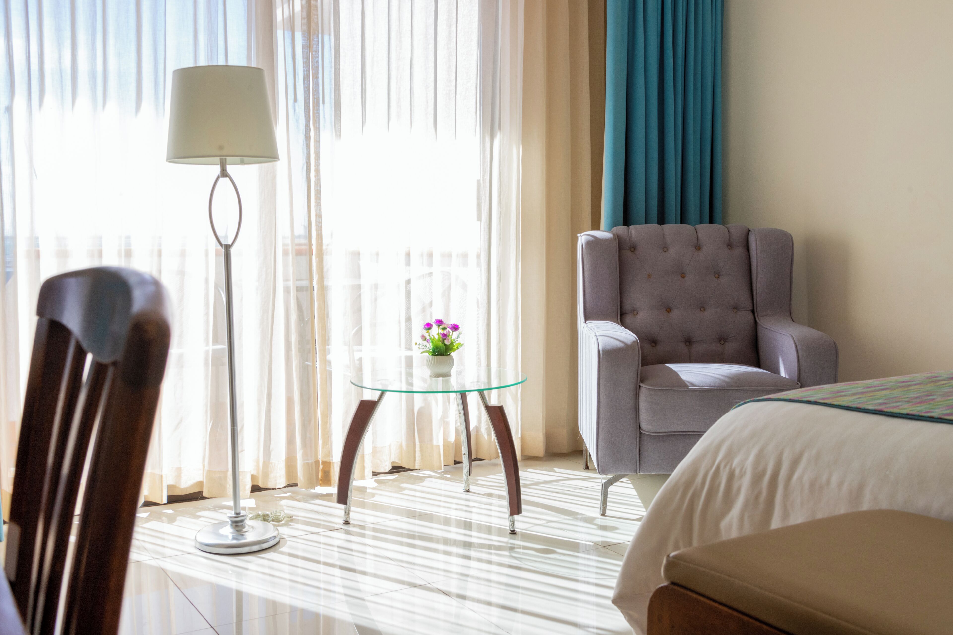 A hotel room with a bed, a grey armchair, a glass table with a vase of flowers, and a floor lamp.