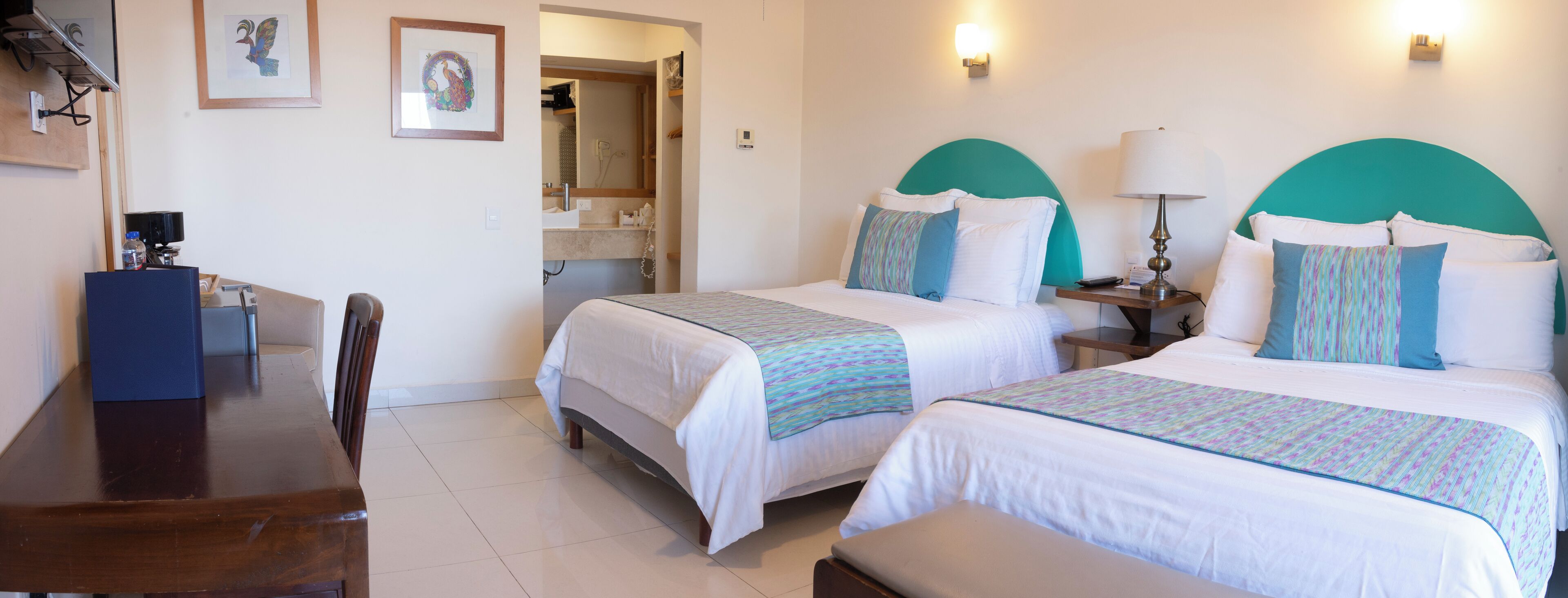A hotel room with two beds, a desk, and a chair. There is a bathroom visible in the background.