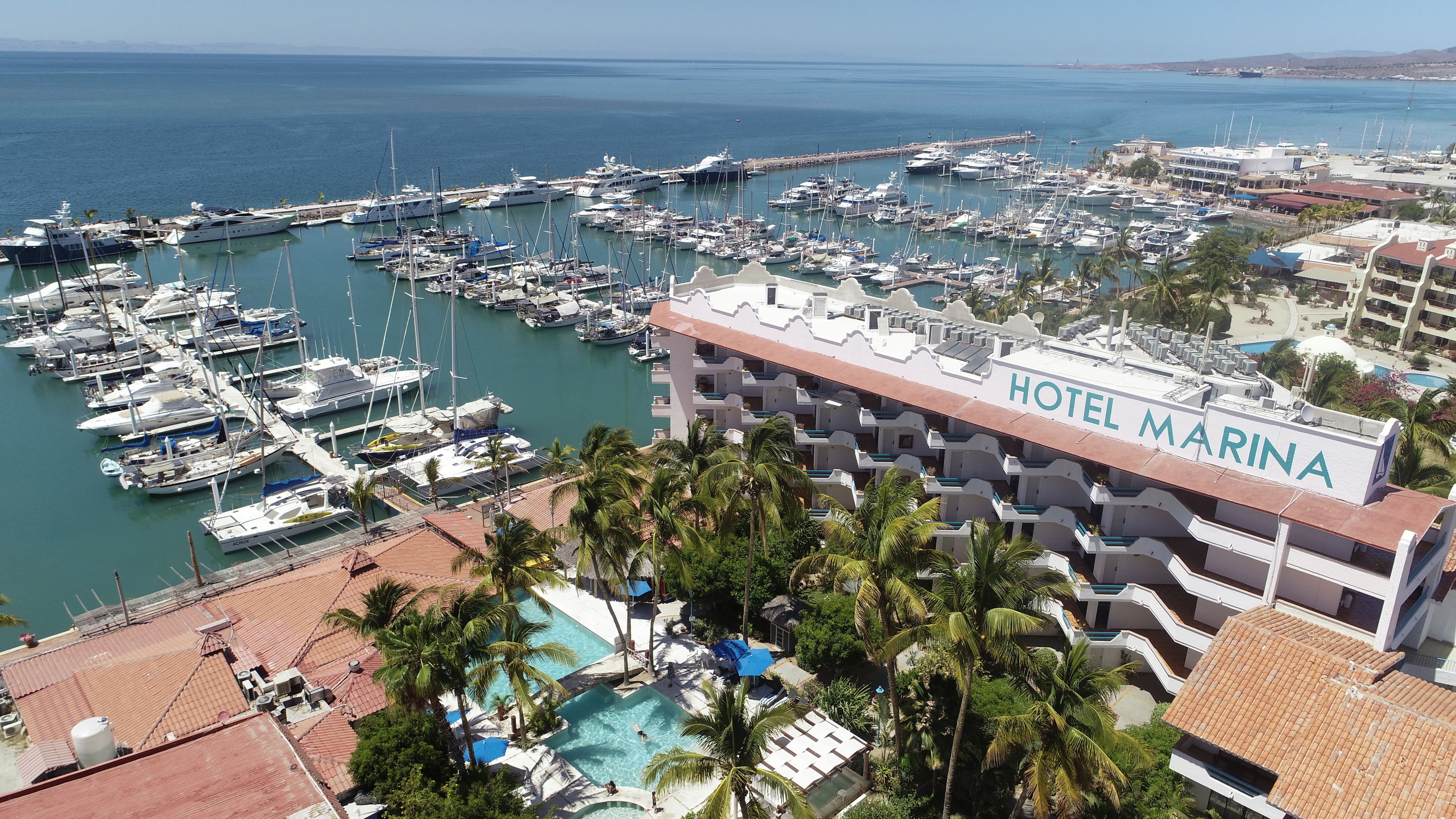 A marina with multiple yachts docked, a hotel named "HOTEL MARINA" with balconies, and a pool area with palm trees.