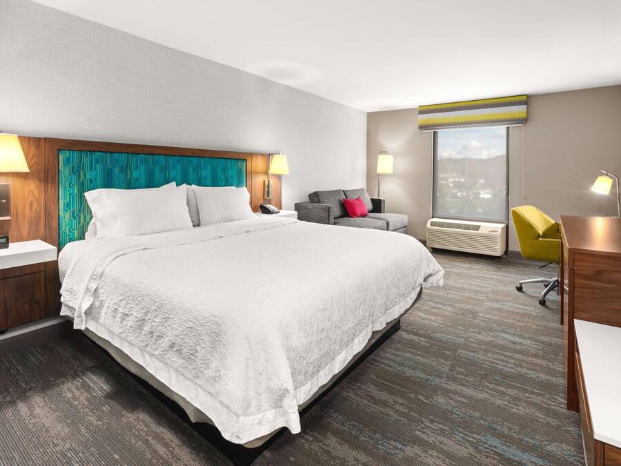Quarto de hotel com uma cama grande, duas mesas de cabeceira com luminárias, uma escrivaninha com cadeira, uma janela com vista e um aparelho de ar condicionado.