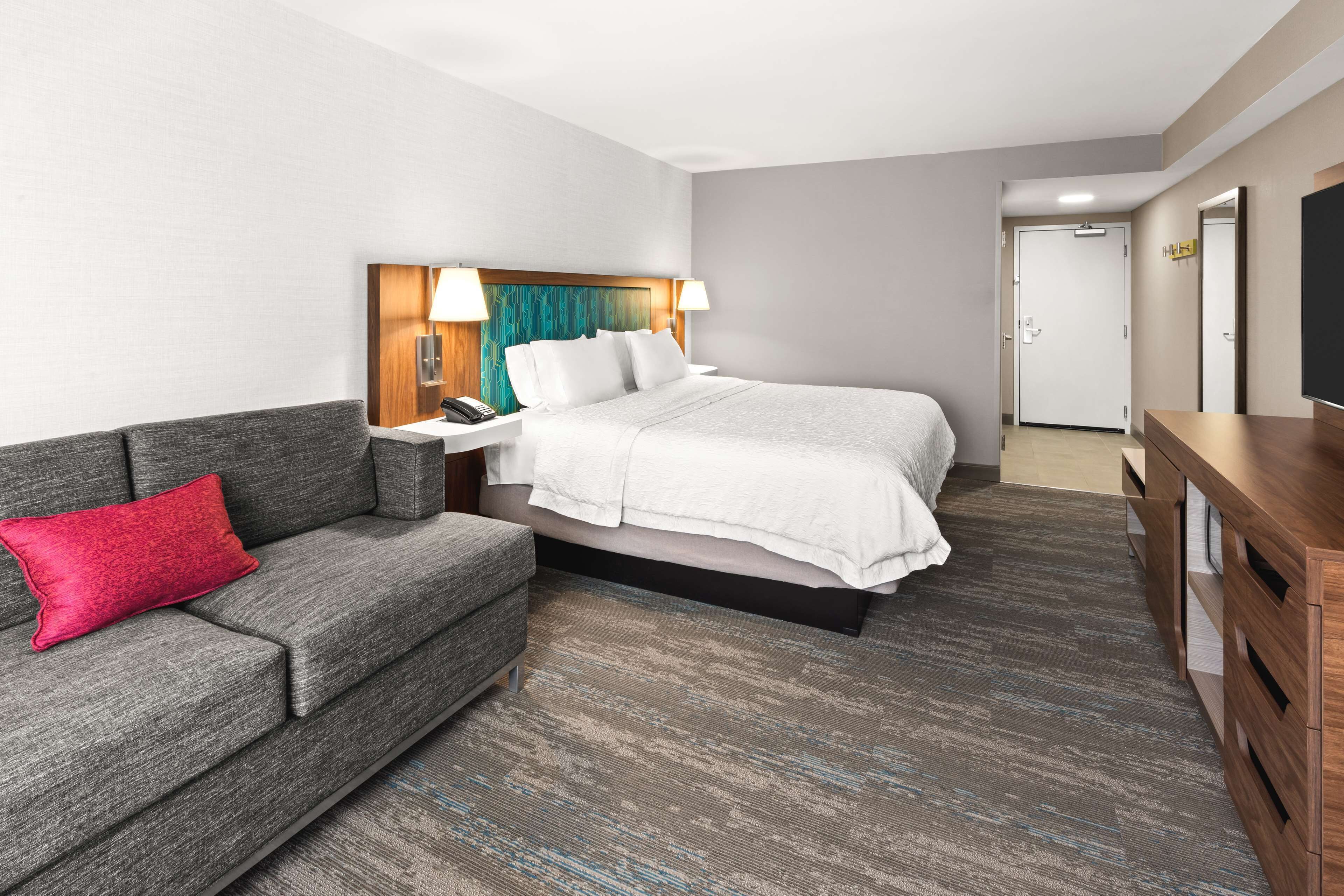A hotel room with a bed, a grey sofa with a red pillow, a wooden nightstand, a wooden dresser, and a television.