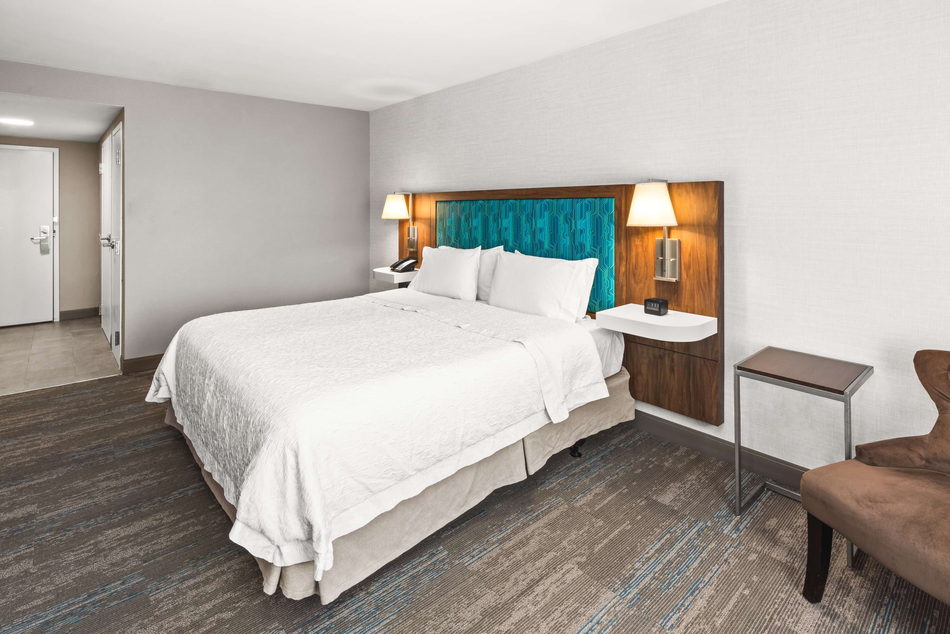 A hotel room with a large bed, two bedside tables with lamps, a chair, and a door leading to another room.