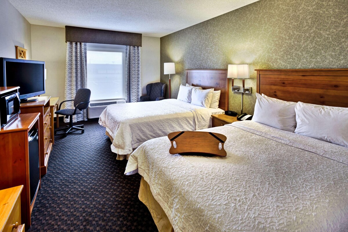 A hotel room with two beds, a desk with a television, a chair, and a window with curtains.