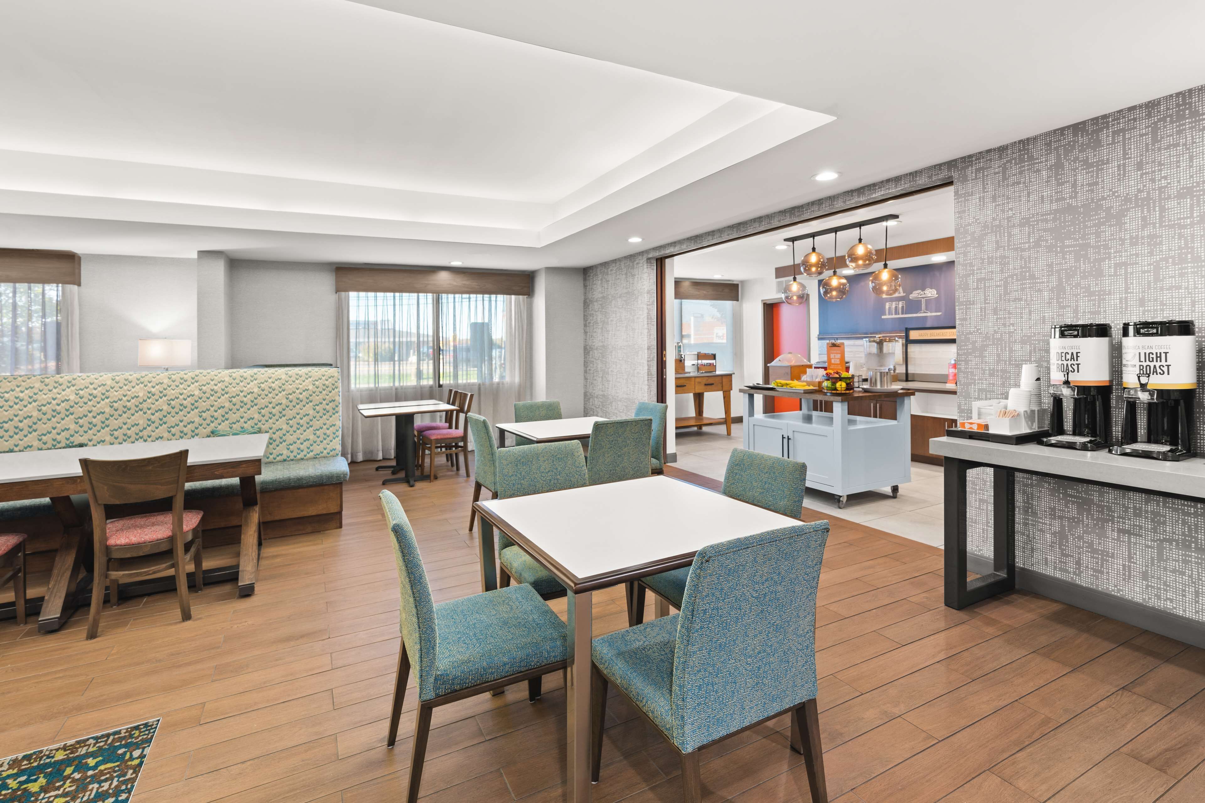 A modern hotel dining area with a long table, several chairs, a sofa, and a kitchen area with coffee machines.