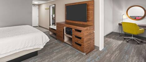 A hotel room with a bed, a wooden entertainment unit with a flat-screen TV, a desk with a chair, and a mirror on the wall.