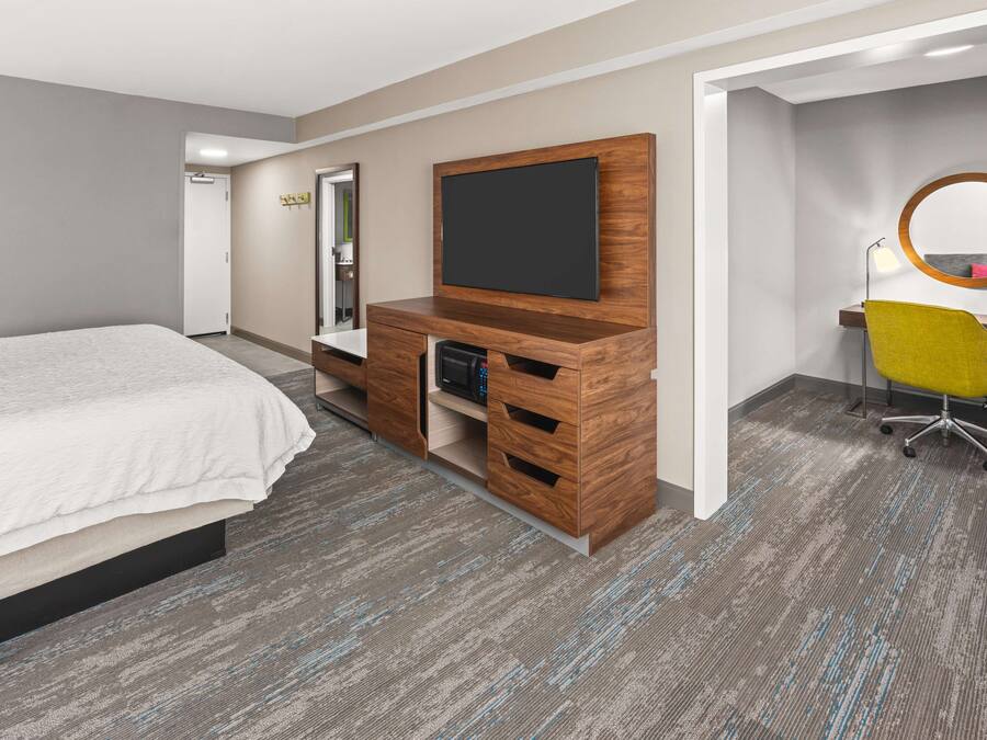 Habitación de hotel con cama, mueble de madera con televisor de pantalla plana, escritorio con silla y espejo en la pared.