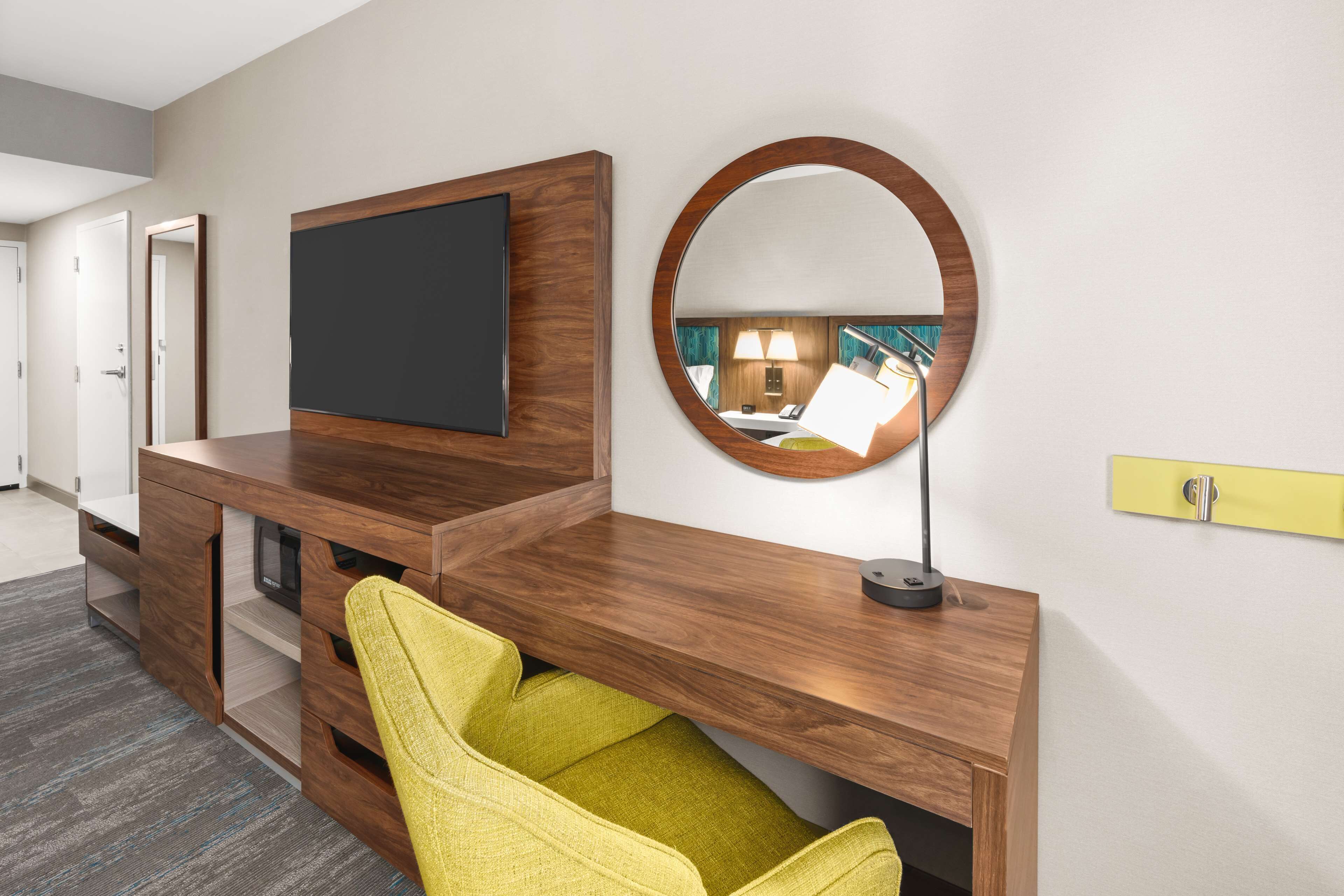 Una habitación de hotel moderna con una mesa consola de madera, un televisor de pantalla plana, un espejo redondo y una silla amarilla.