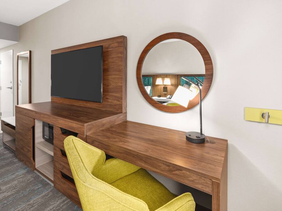 Una habitación de hotel moderna con una mesa consola de madera, un televisor de pantalla plana, un espejo redondo y una silla amarilla.