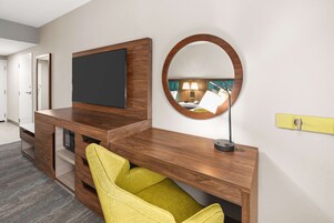 Una habitación de hotel moderna con una mesa consola de madera, un televisor de pantalla plana, un espejo redondo y una silla amarilla.