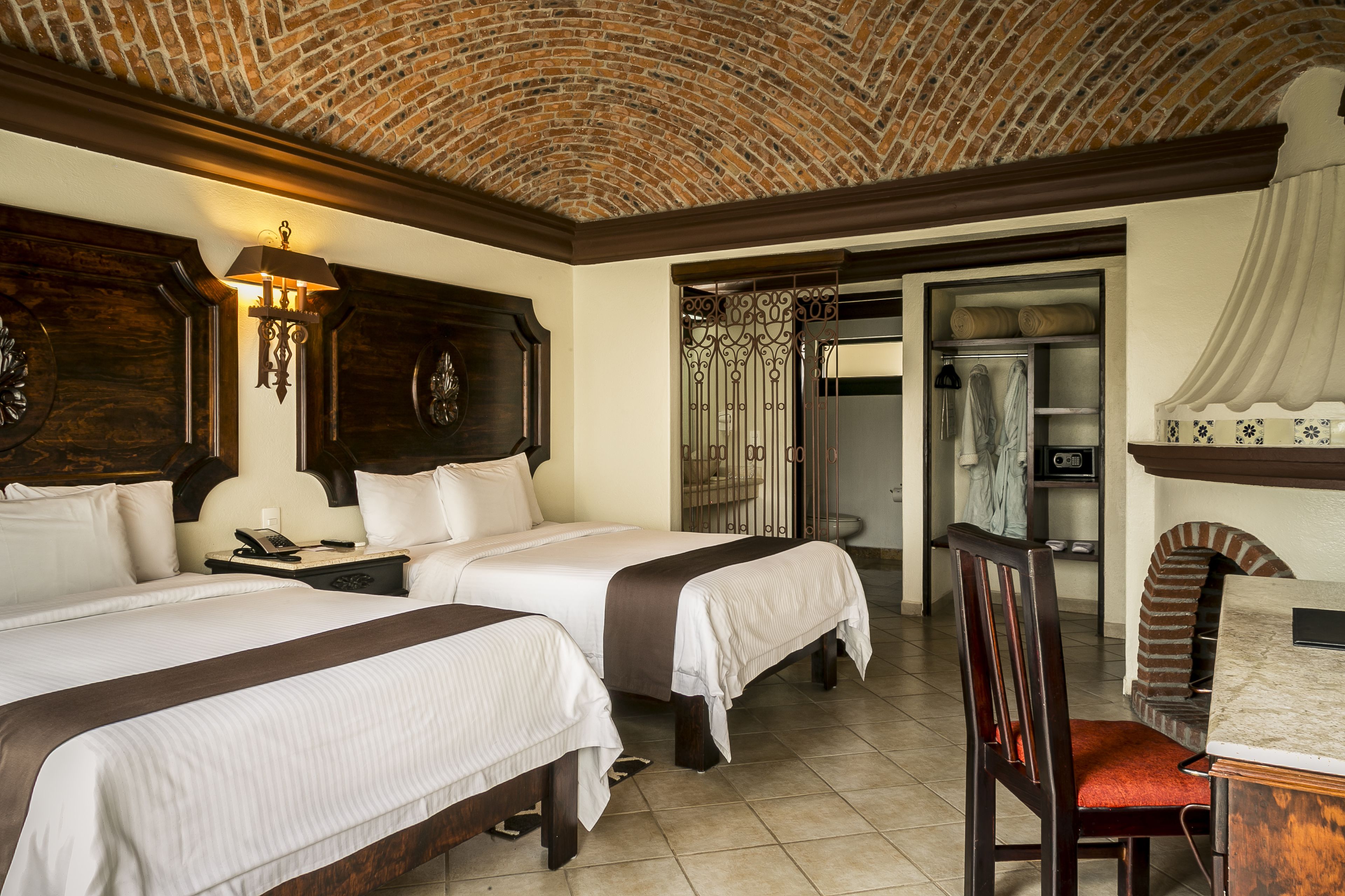 Habitación de hotel con dos camas, cabecero de madera, techo de ladrillo, una mesita con una silla y un baño visible a través de una puerta.