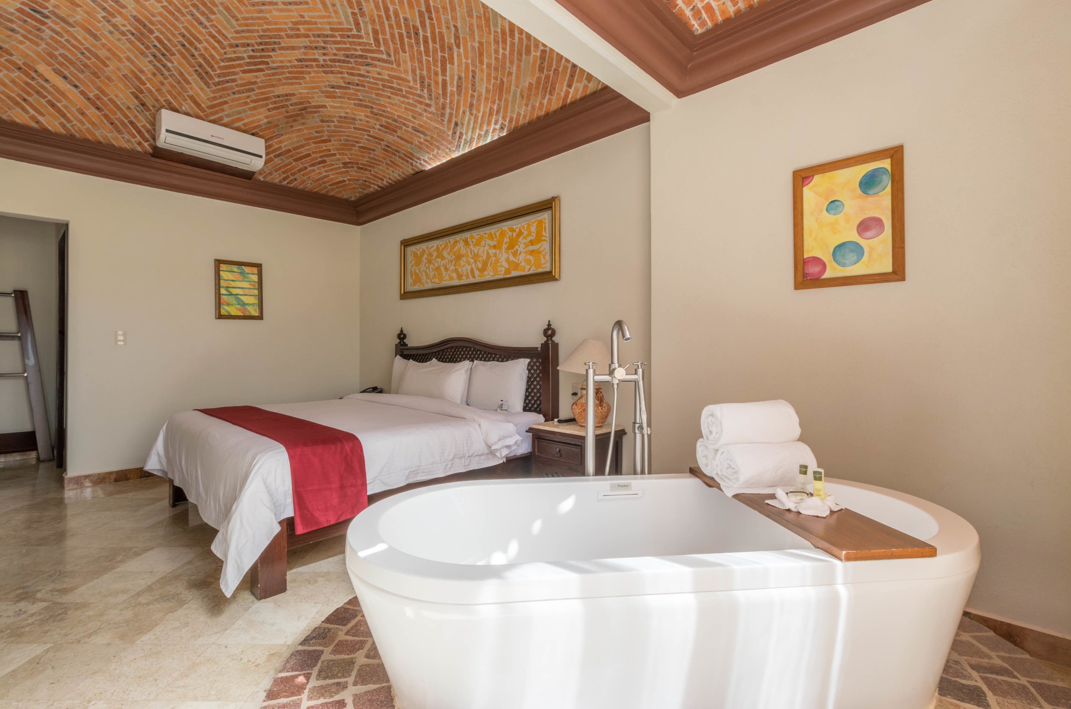 A hotel room with a large white bathtub, a bed with a red blanket, a wooden headboard, a nightstand with a lamp, and a painting on the wall.