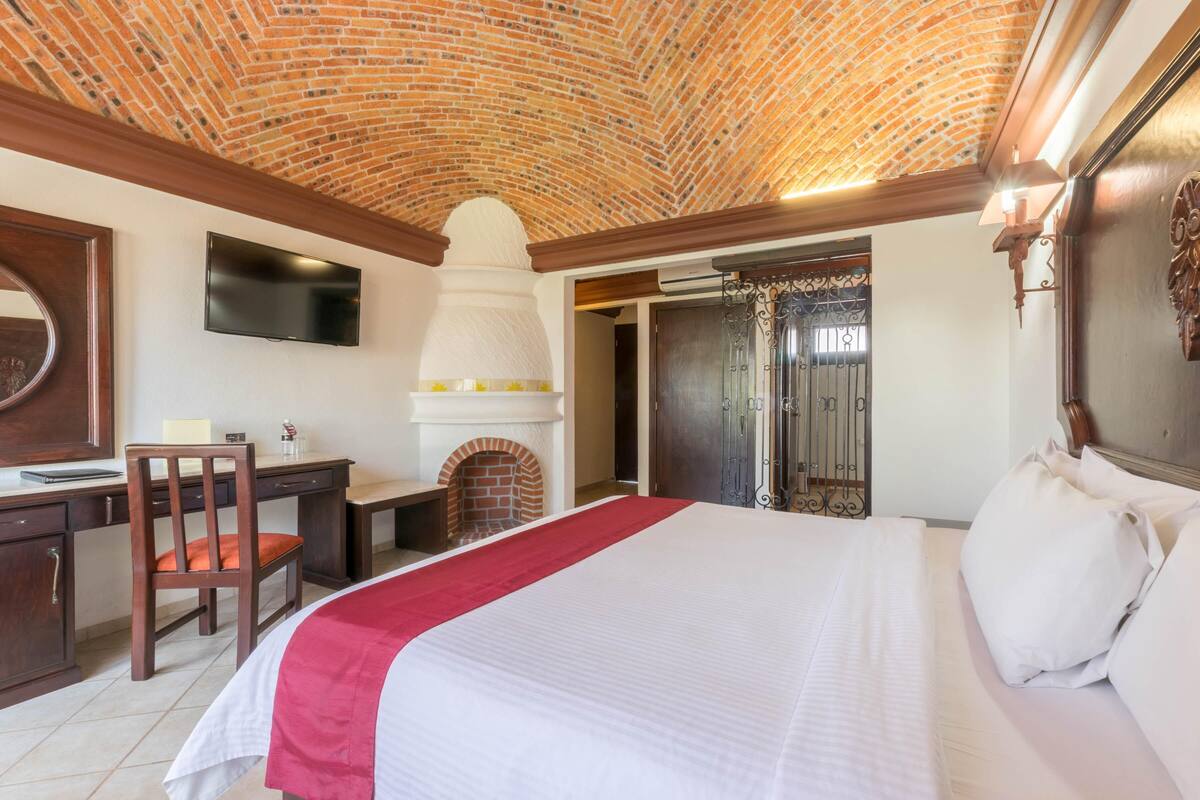 Ein Hotelzimmer mit Ziegeldecke, Kamin, Schreibtisch mit Stuhl, Bett mit weißer Bettwäsche und einem an der Wand montierten Fernseher.