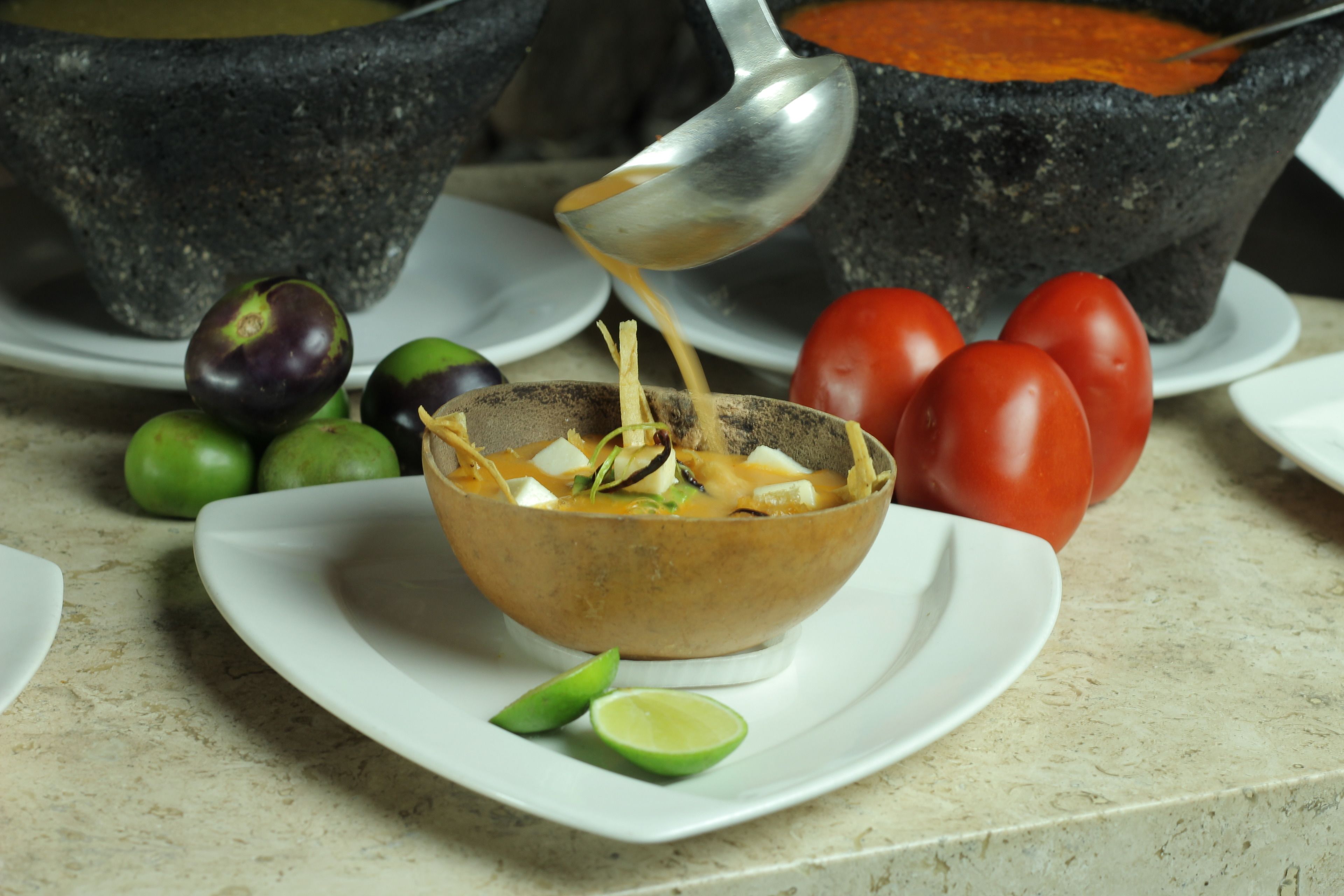 Un tazón de sopa con una cucharada levantándose, rodeado de tomates, limones y un mortero.