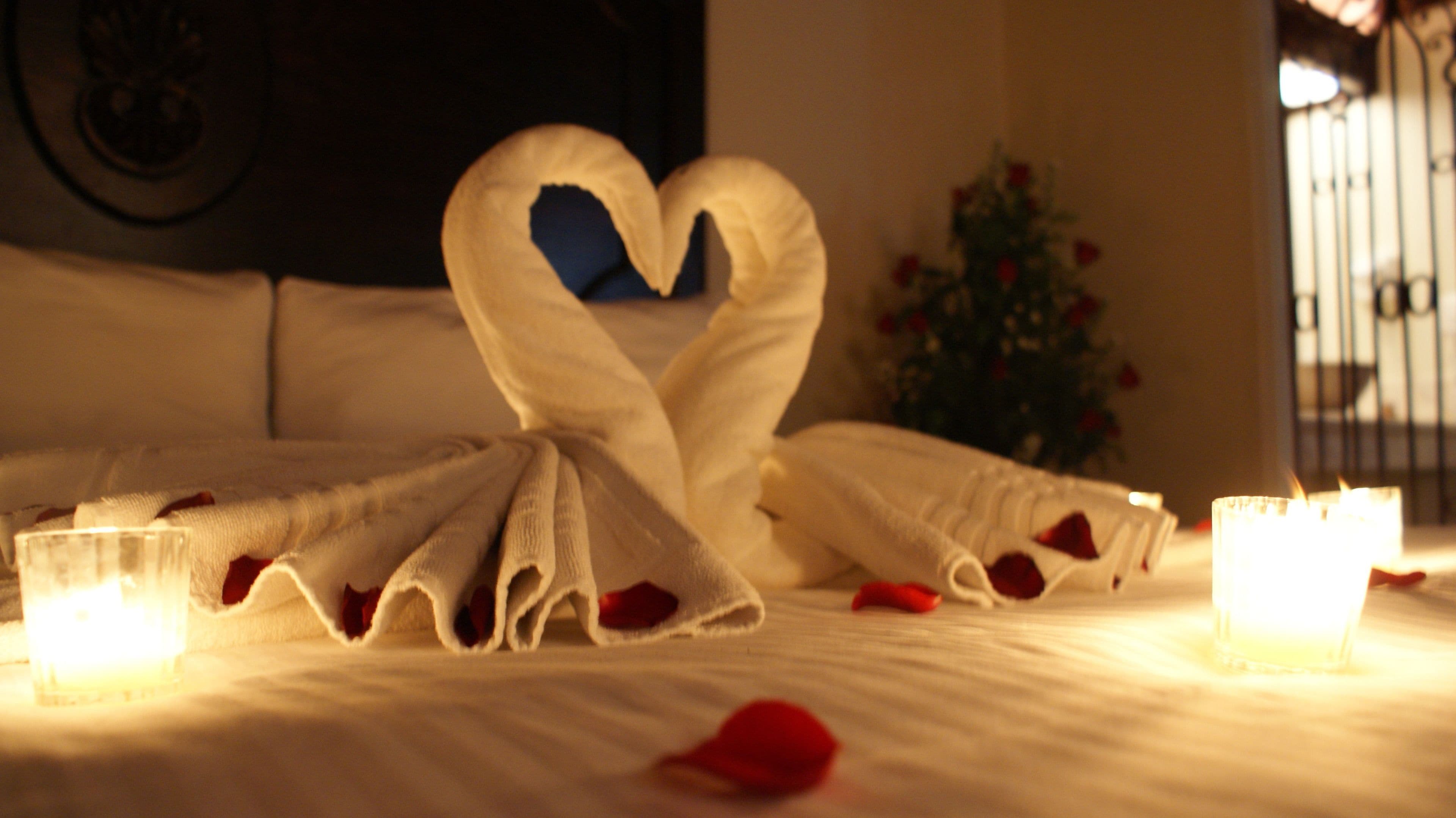 Una cama con una toalla dispuesta en forma de corazón, velas y pétalos de rosa.