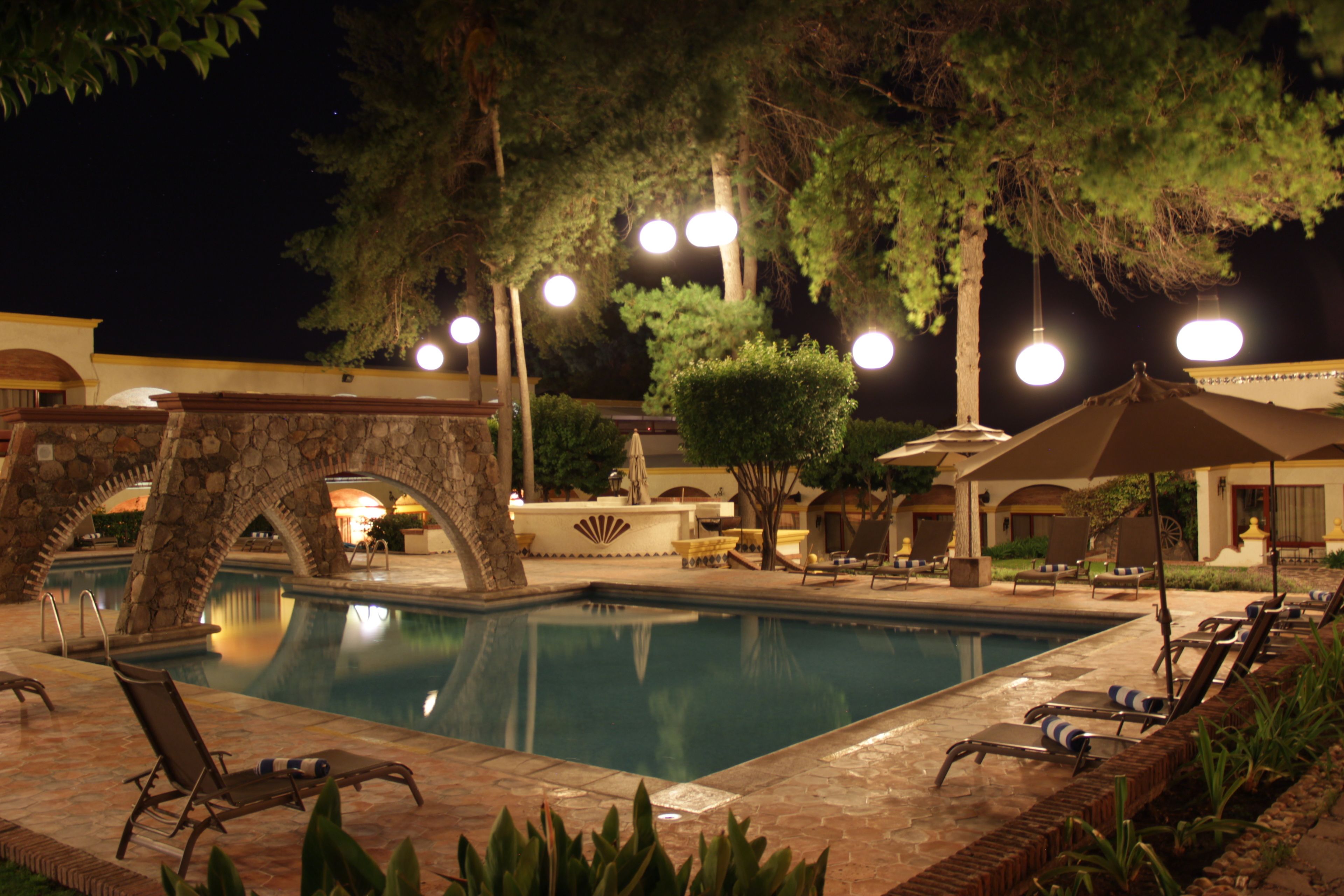 Zona de piscina nocturna con sillones y sombrillas, rodeada de árboles y arquitectura de piedra.