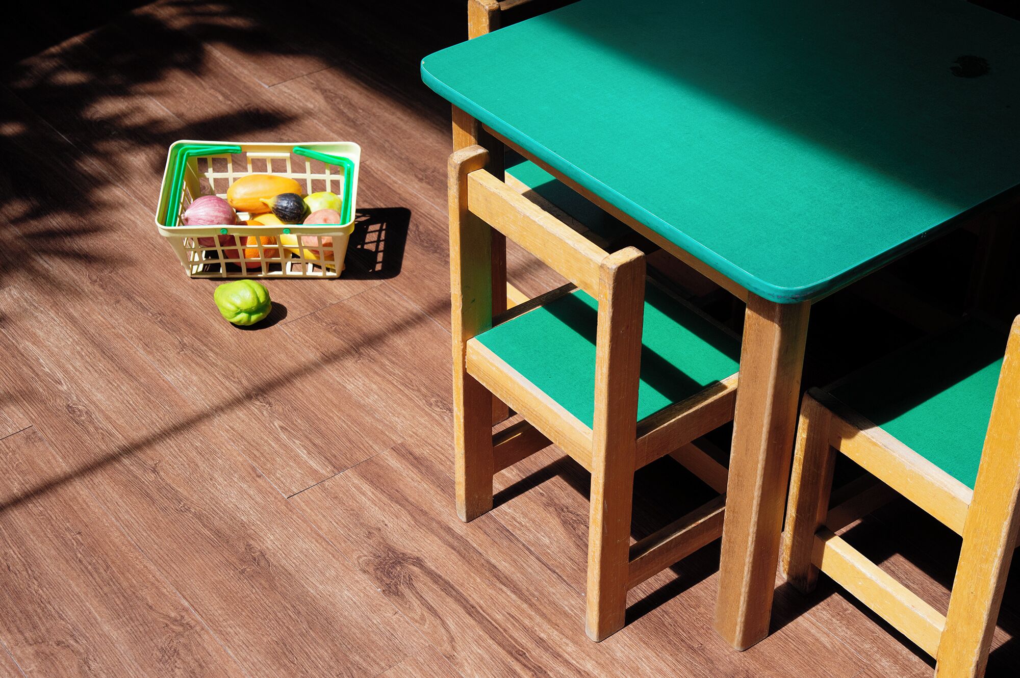 Una mesa de madera con cubierta verde y sillas a juego, una cesta de frutas y una pelota verde sobre un piso de madera.