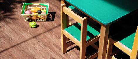 Una mesa de madera con cubierta verde y sillas a juego, una cesta de frutas y una pelota verde sobre un piso de madera.
