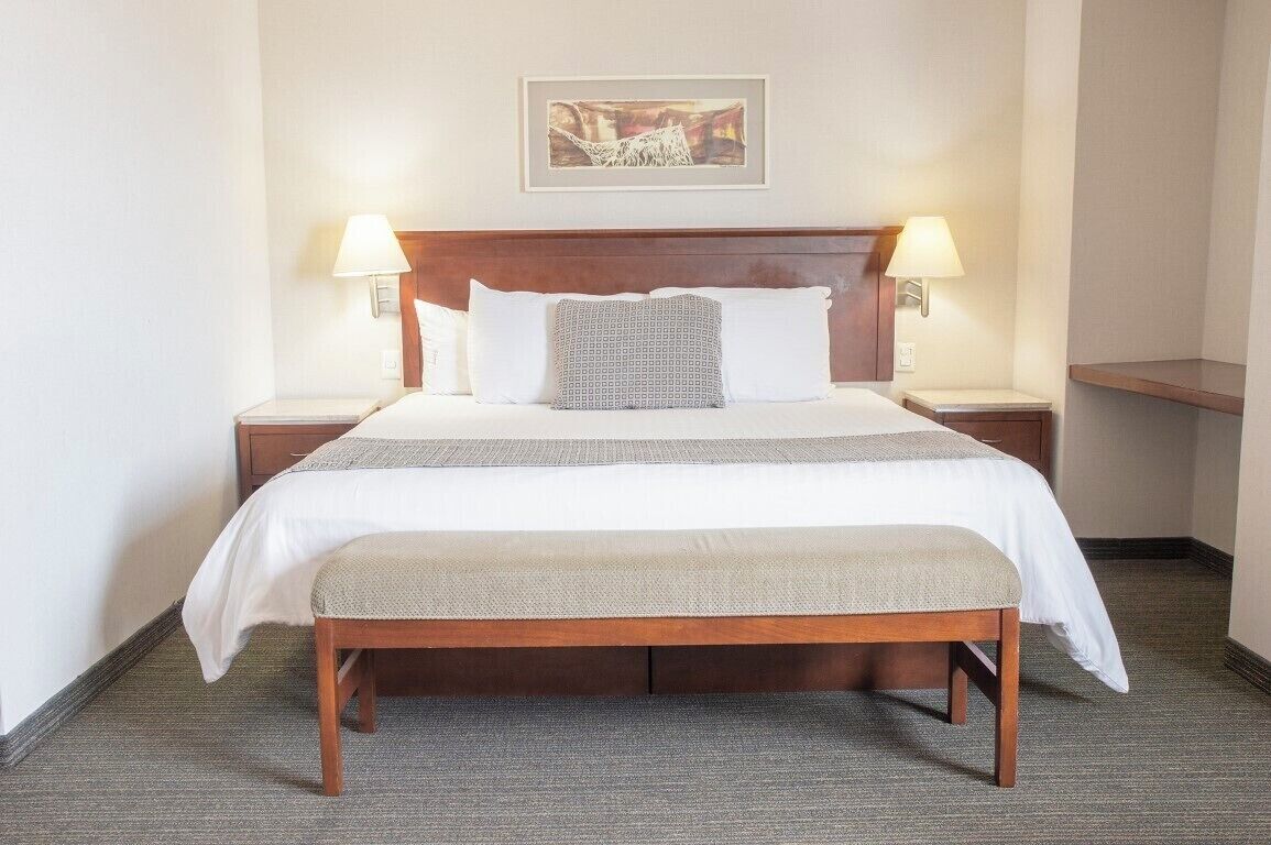 Quarto de hotel com cama de madeira, roupa de cama branca, um banco no rodapé da cama, duas luminárias de cabeceira e um quadro emoldurado na parede.