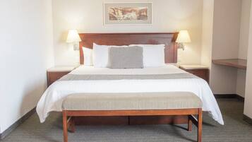 Quarto de hotel com cama de madeira, roupa de cama branca, um banco no rodapé da cama, duas luminárias de cabeceira e um quadro emoldurado na parede.