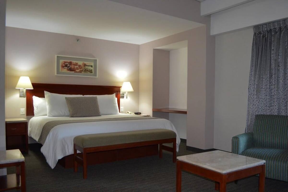 A hotel room with a bed, bedside lamps, a bench, a small table, and a chair.
