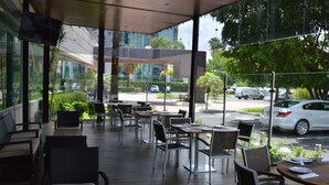 Uma área de café ao ar livre com mesas e cadeiras, janelas amplas e vista para a rua.