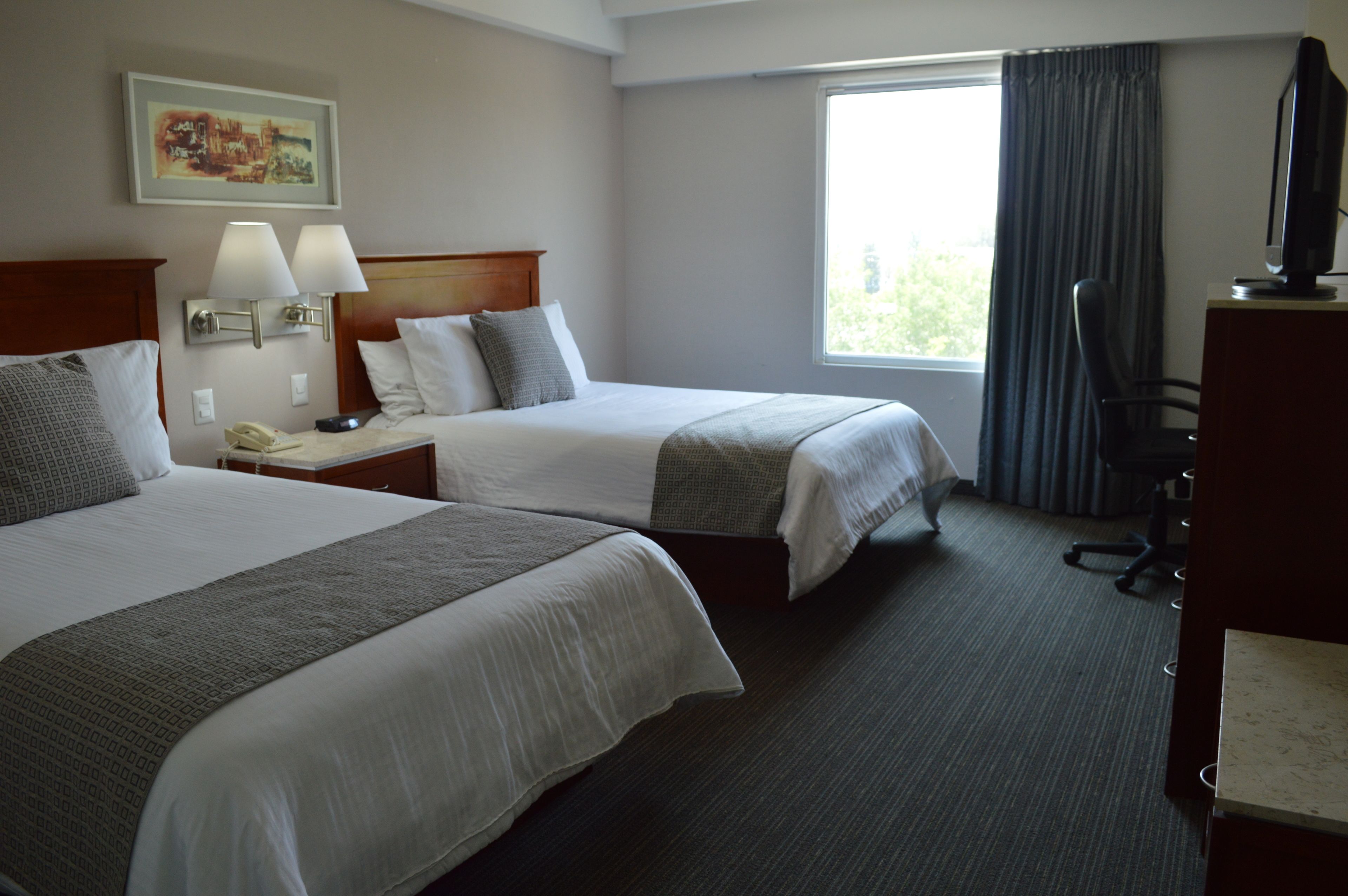 Habitación de hotel con dos camas, un escritorio con computadora, una lámpara y una ventana con cortinas.