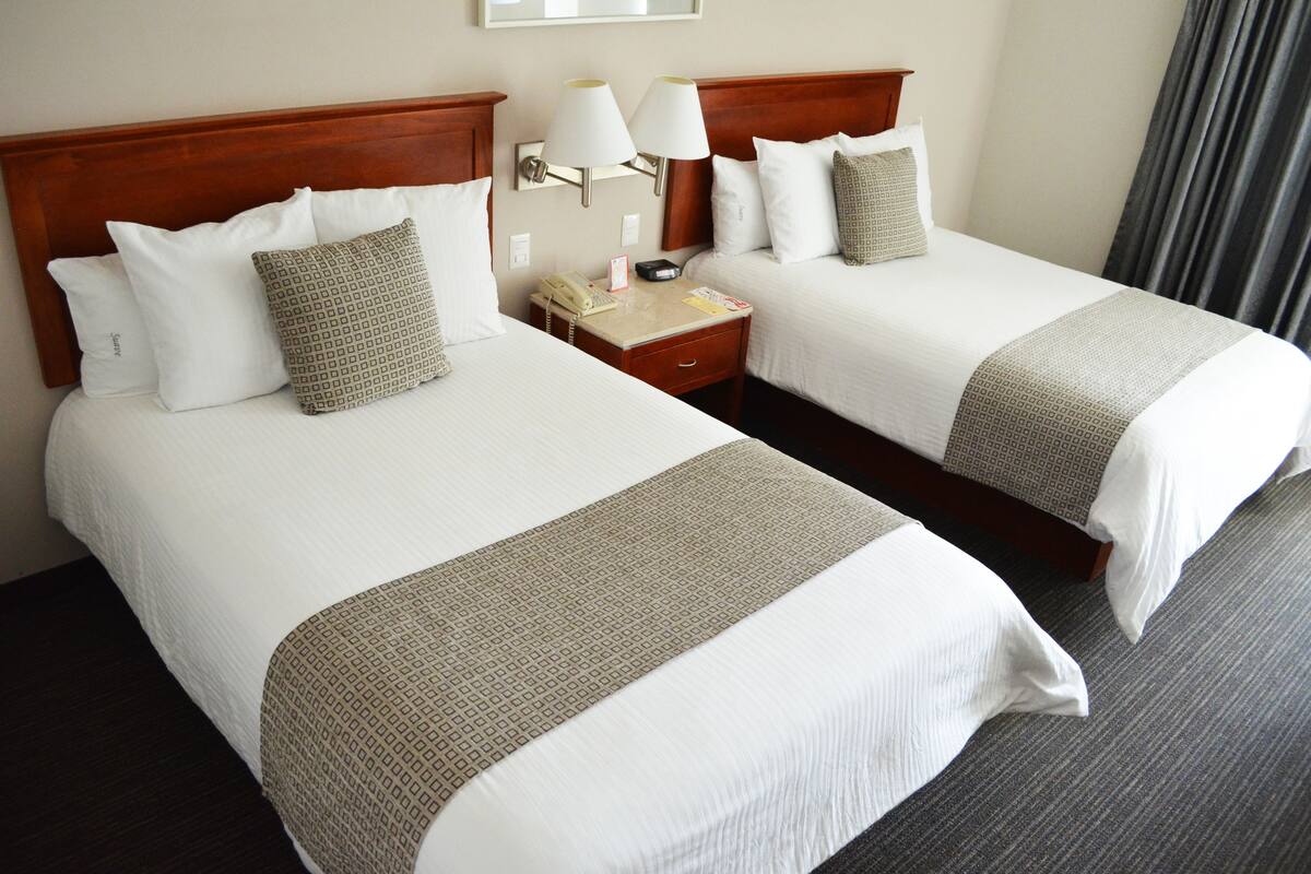 Two adjacent hotel beds with white linens, a wooden headboard, a nightstand with a lamp, and a wall-mounted light fixture.