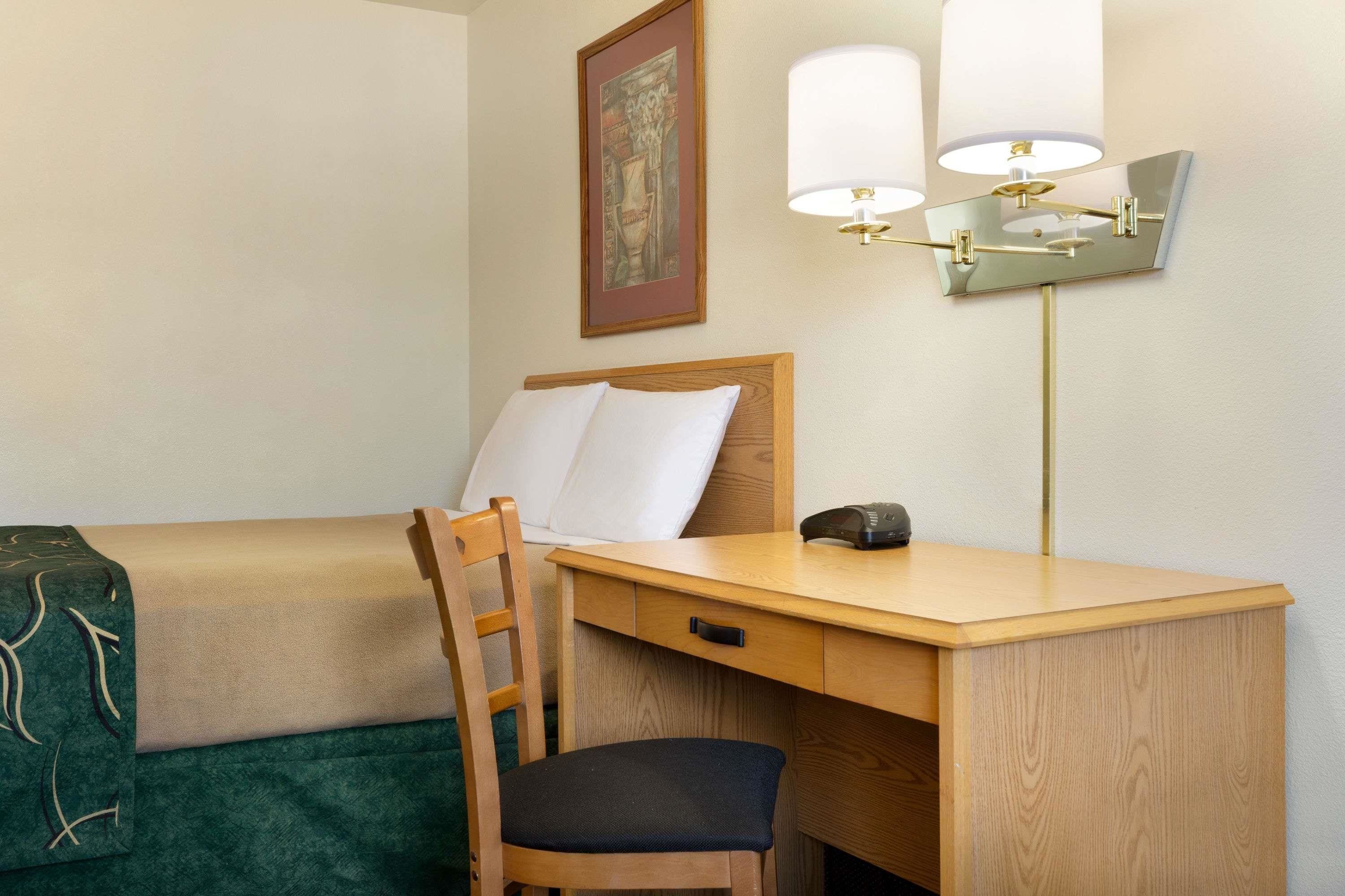 Habitación de hotel con un escritorio de madera, una silla, una cama con manta verde y una lámpara de pared.